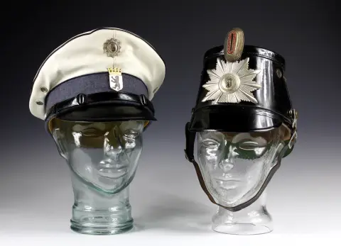 Kuriositeter – To hodeplagg fra Berlins politi. Politihjelm "Tschako" 1945-1968, hvit politihatt 1954-1961. Løse på hatteholdere av glass.