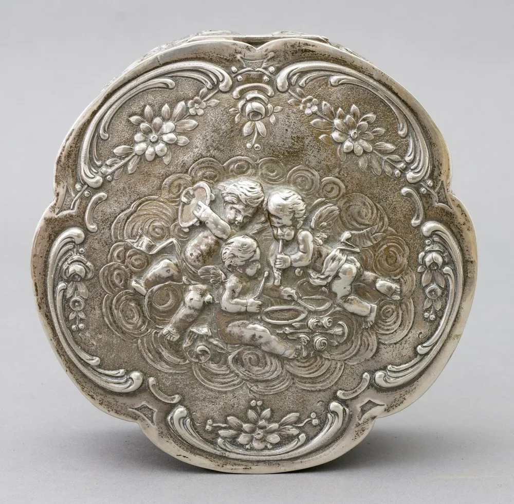 Box, 800 silver, German, Wilhelm Weinranck & Fritz Schmidt, Hanau, 3rd Rococo, 1900 - 1950 decor of flowers, rose blossoms, rocailles and numerous, some music-making putti. Hinged lid. D 16 cm, H 6.3 cm, weight 400 g. Marked on the base: 800, crescent, crown and maker's mark WSH in heart.